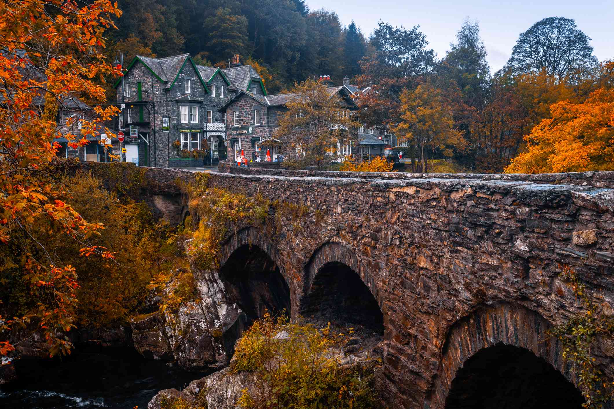 在威尔士的斯诺登尼亚，一座被明亮的橙色树木环绕的古老石桥和两所背景是绿色和金色树木的小屋