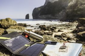 Portable solar panel charging a laptop near the ocean.