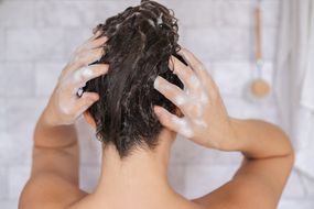 女人的白色瓷砖淋浴用手擦头发的洗发水”width=