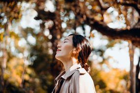 年轻的亚洲女子的画像在公园里散步,享受阳光的温暖美丽的秋天的一天在户外和呼吸新鲜的空气,闭上眼睛。放松在枫树下的性质