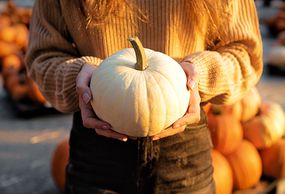 woman in brown sweater holds white pumpkin in front of pumpkin patch