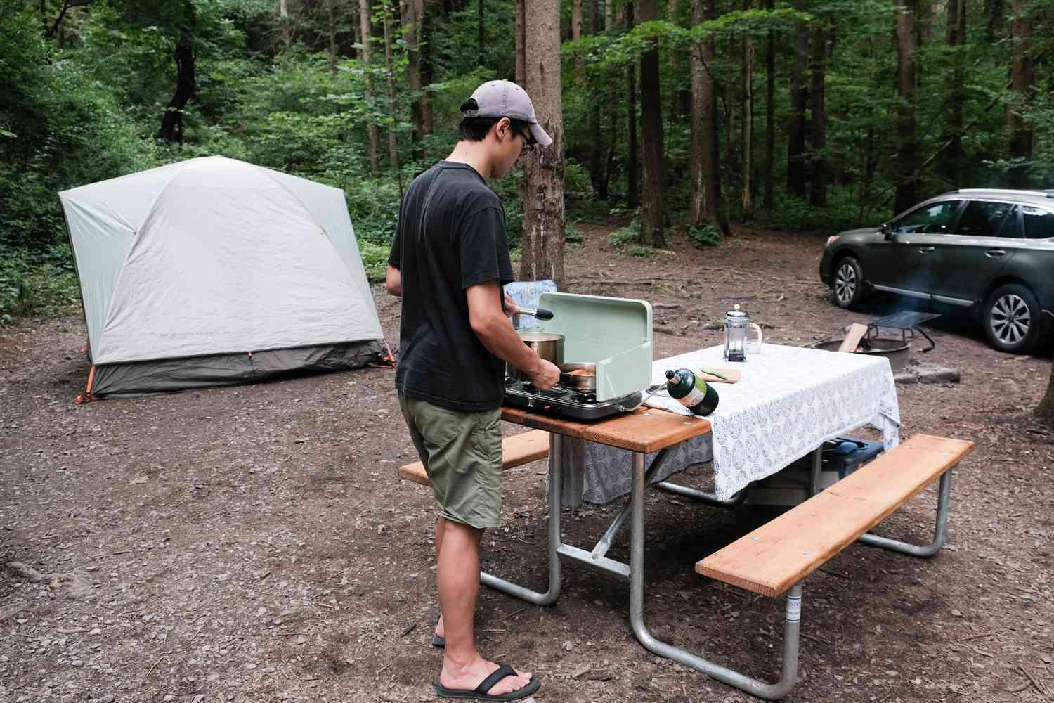 一名男子在野餐桌上的野营炉上做饭，附近有帐篷和汽车