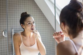 成年妇女在浴室镜子里刷牙“width=