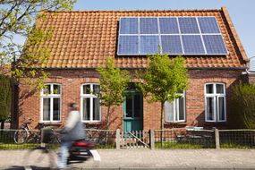 在房子前面的人骑自行车有太阳电池板的“width=