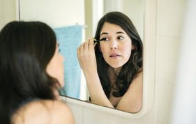 一名深色的女人将睫毛膏施到镜子中的眼睛。“width=