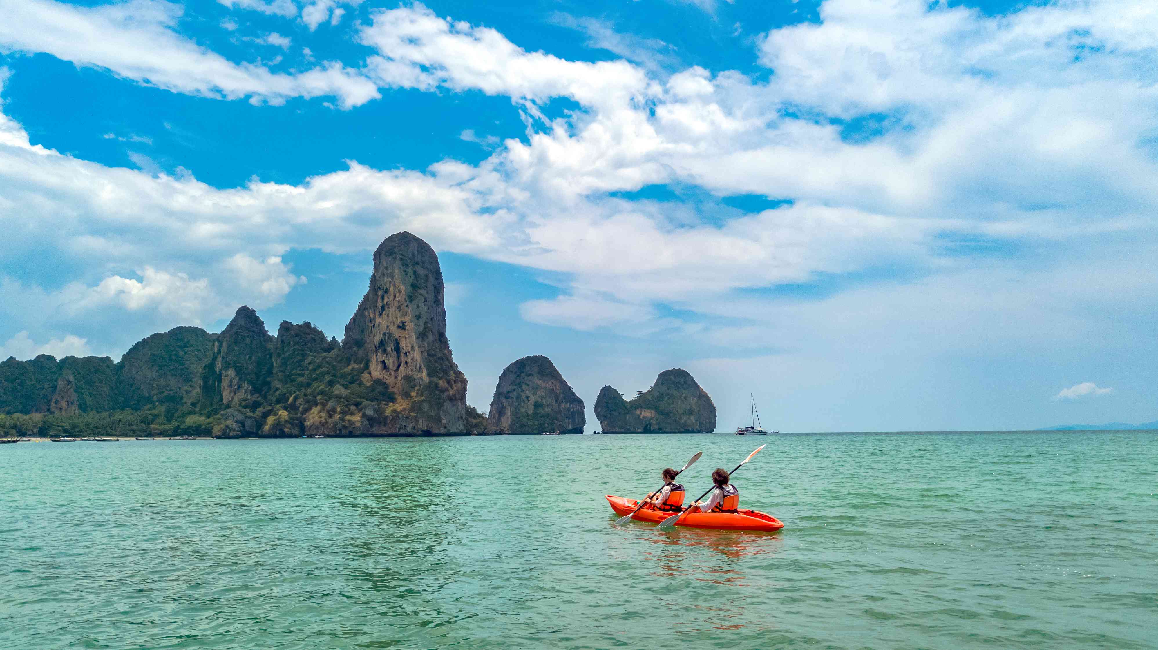 在泰国甲米，两个人乘坐红色皮艇在蓝绿色的水中划向一处岩层。