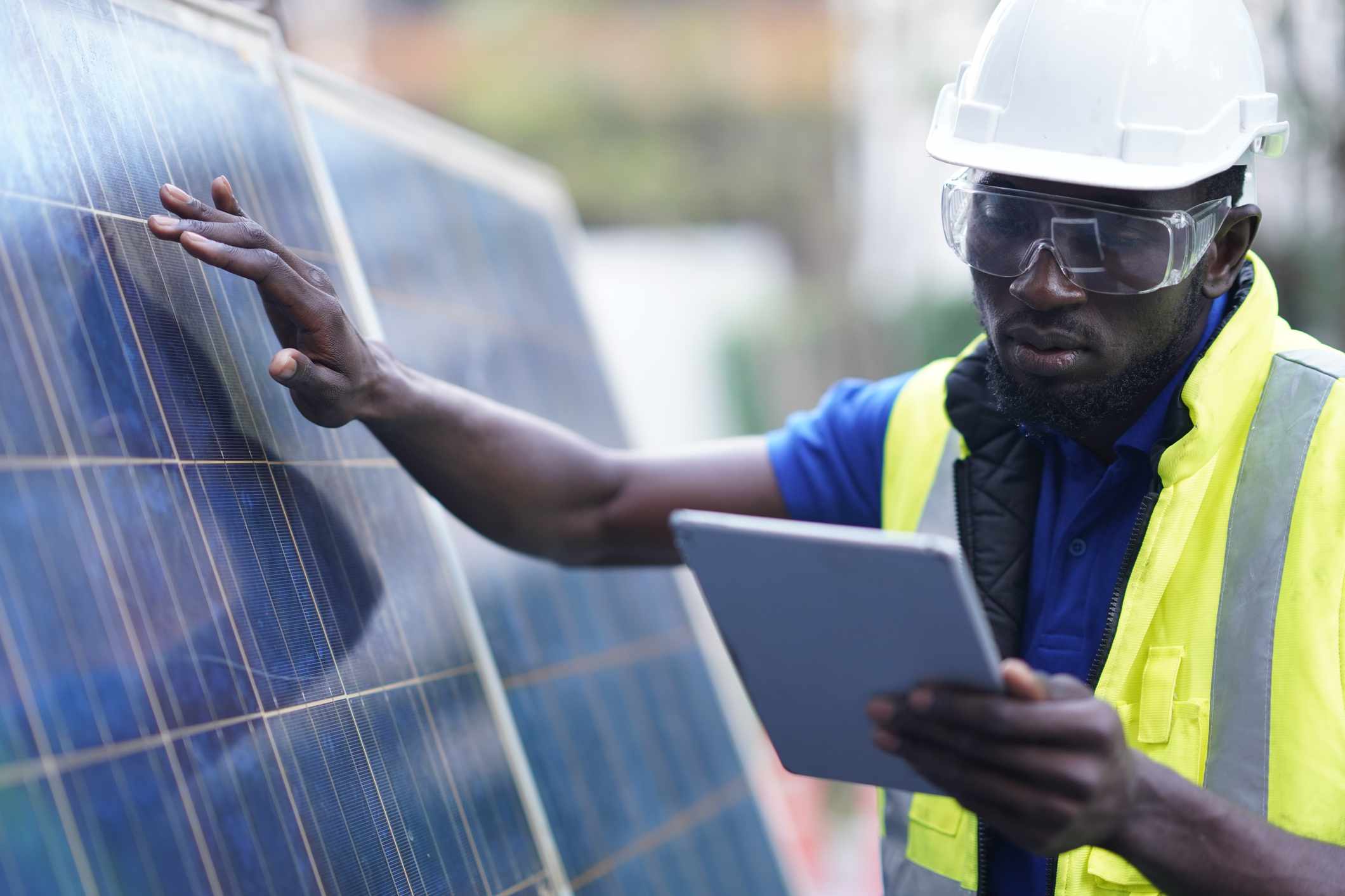 一名非洲裔太阳能系统工程师戴着护目镜和白色安全帽，正在分析太阳能电池板的能源效率。＂width=