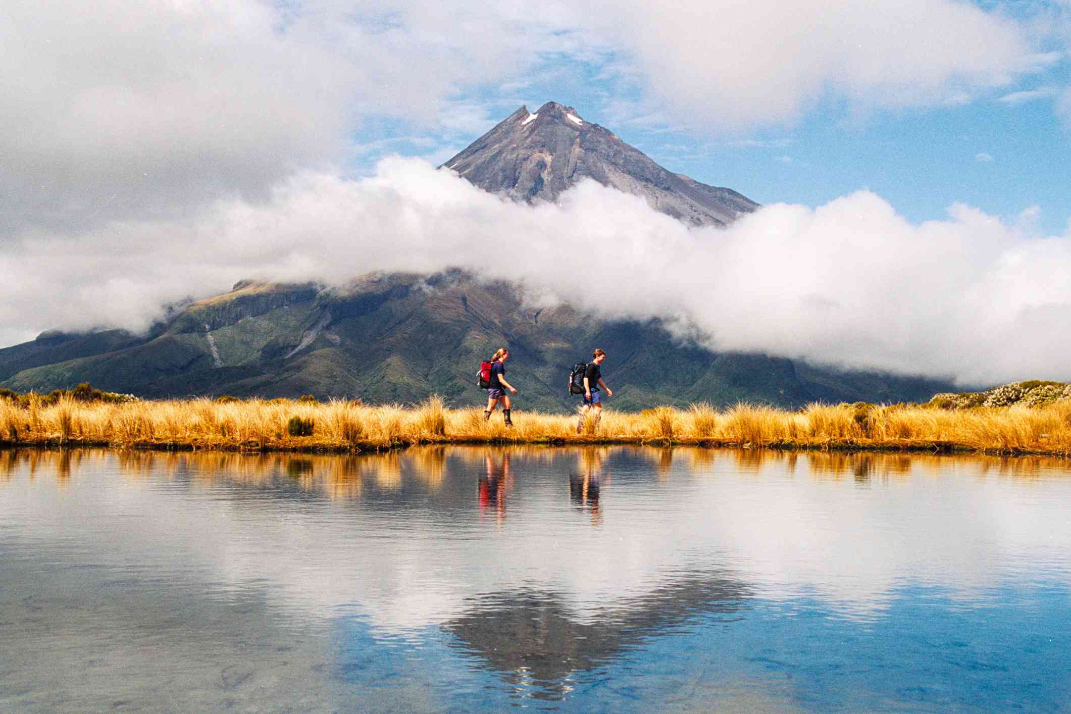 两个徒步旅行者走在火山前反光的湖上＂width=