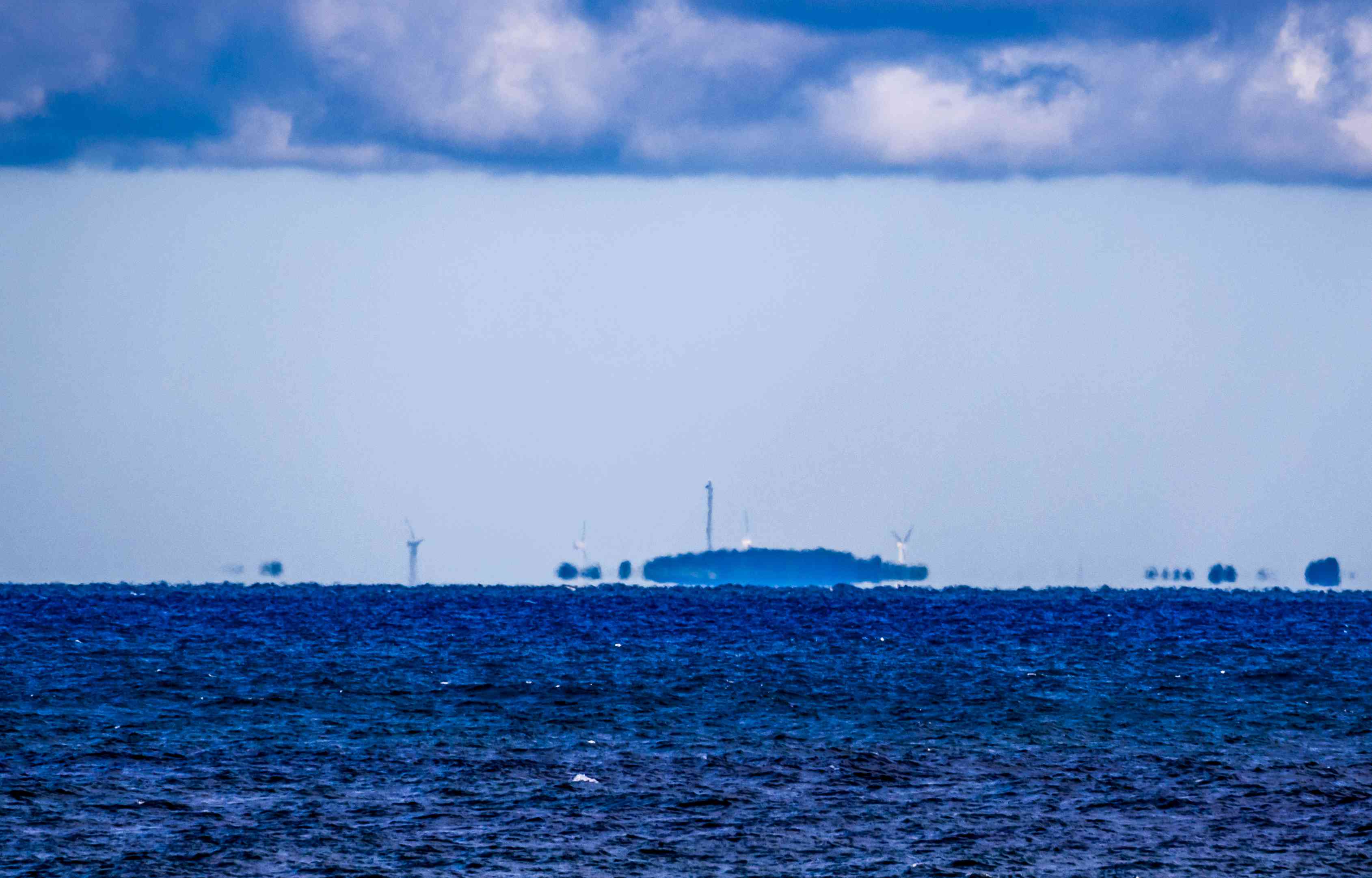从Fata Morgana的海洋上俯瞰海岸线的海市蜃楼，上面有风力涡轮机，天空多云，有一些降水