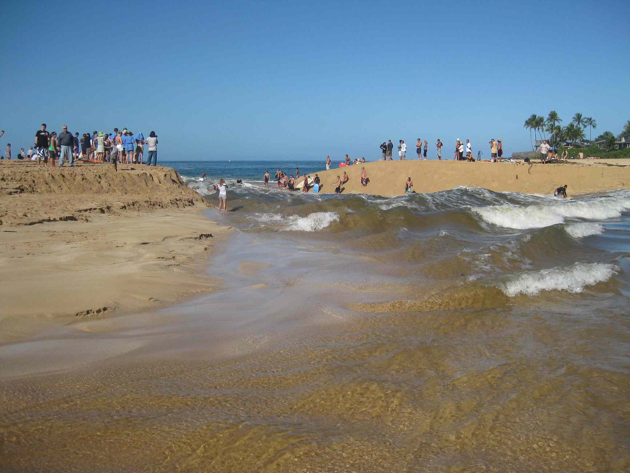 海浪突破了威美亚河的嘴，在海浪的两侧，挤满了沙丘，观察者看着在明亮的蓝天下的水中看着一些冲浪者