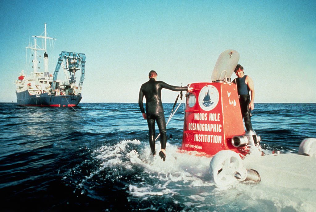一张旧照片，一艘潜水器从海洋中浮出水面，两个穿着潜水服的人站在上面，背景是一艘船。＂width=