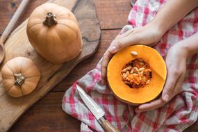 holds cup a pumpkin cut in half to reveal seeds for saving with knife and cutting board