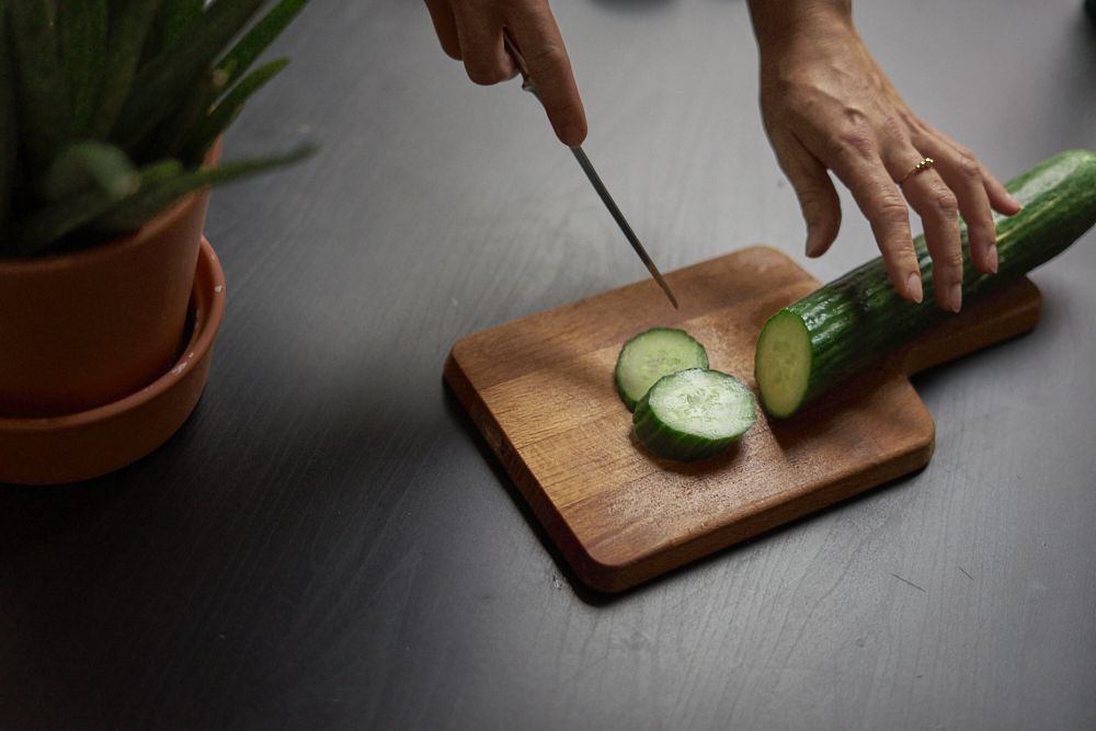 手在木制切菜板上切成绿色黄瓜，切成薄片“width=