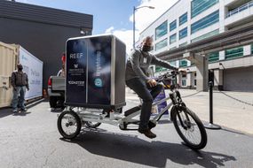 一个人在一个城市货运实验室e-cargo自行车戴着口罩。”width=
