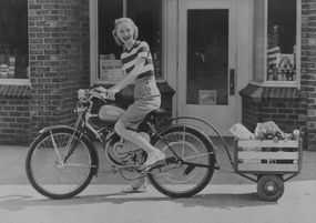 一个女人骑着一辆带着货物拖车的自行车的老式照片＂width=