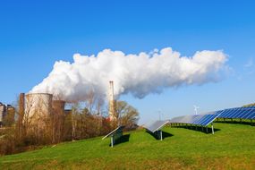 Solar panels on a grassy hillside with a fossil fuel power plant and a single wind turbine in the background