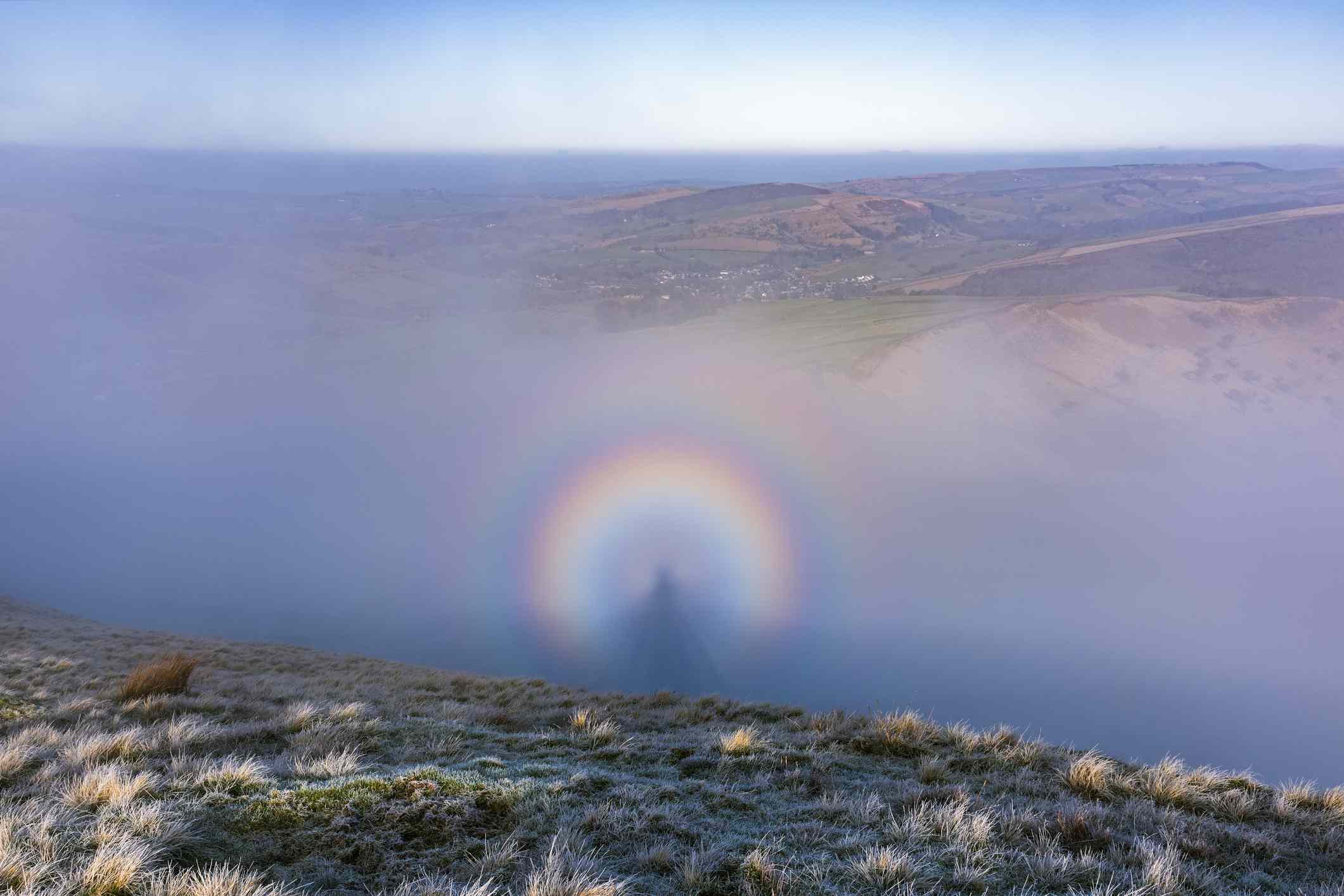 一个观察者被放大的影子投射在太阳对面的云层上，被称为“布罗肯幽灵”，看起来像一个人被彩虹包围着，上面有山脉和蓝白色的天空