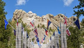 在拉什莫尔山(Mount Rushmore)前的混凝土柱上悬挂着56个州、地区、联邦和地区的旗帜，周围是高大的绿色树木，蓝天下是白色的圆顶