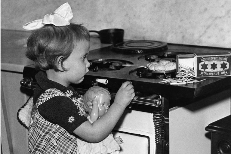一个小女孩抱着娃娃，试图用火柴点燃燃气炉，以烹饪饼干，华盛顿特区，大约1932年。“class=