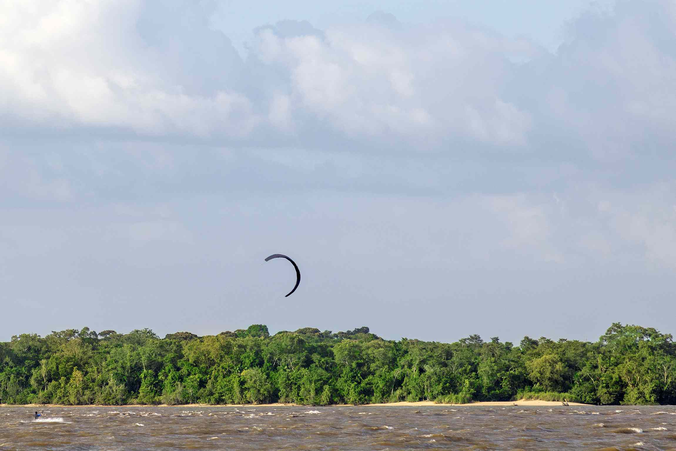 深色亚马逊河的风筝冲浪者，在蓝天下面的树木茂密的树林上空，有几块白云
