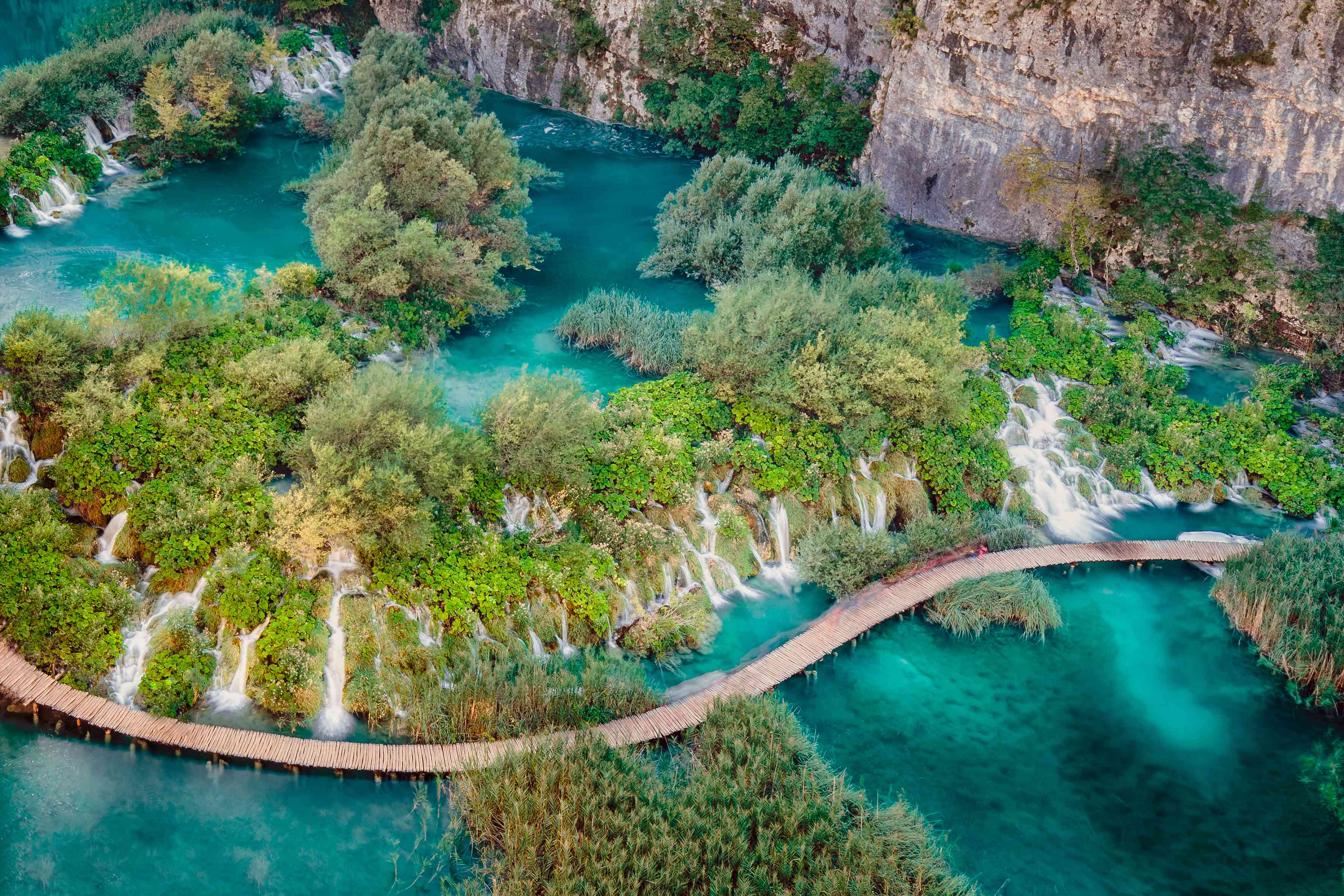 鸟瞰层叠的Plitvice瀑布，瀑布被绿色的树木、山脉和清澈的蓝绿色的水包围＂width=