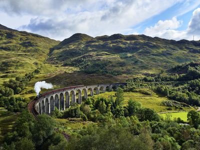 格伦芬南高架桥上的苏格兰高地郁郁葱葱的绿色山丘鸟瞰图，詹姆斯二世党人蒸汽火车在蓝天白云下经过