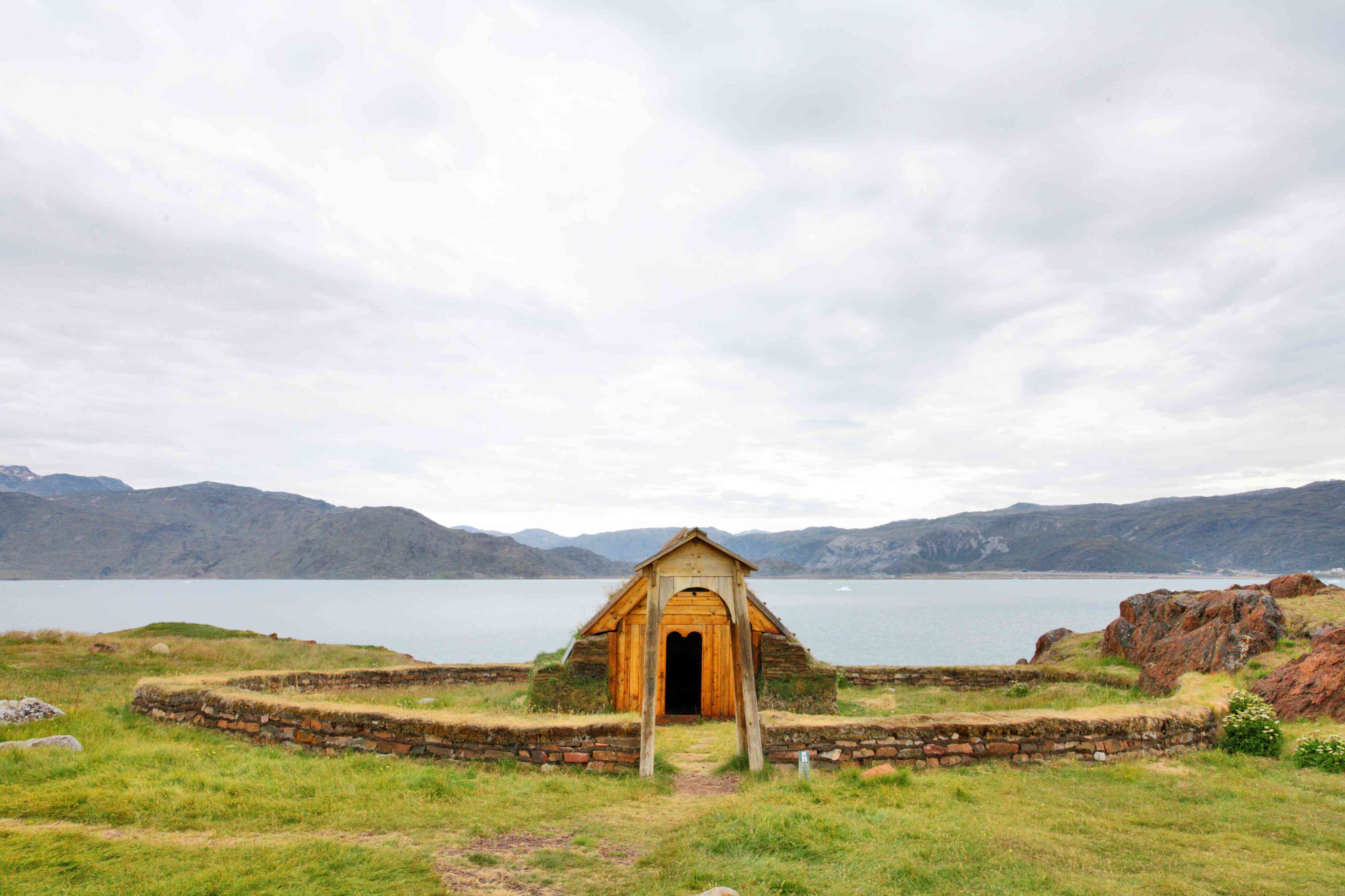 孤独的橙色小屋，低矮的圆砖墙围着它，后面是水和山＂width=